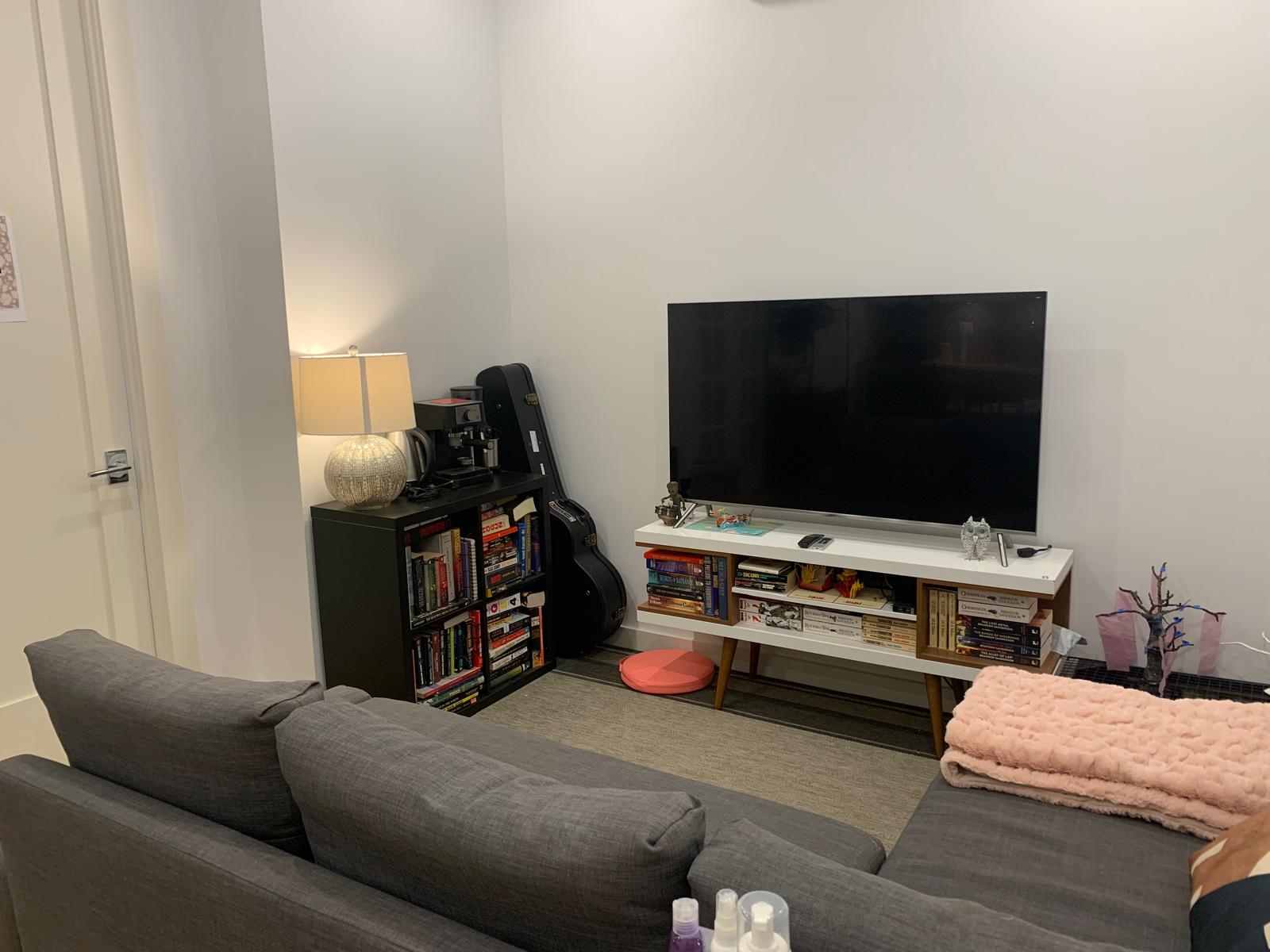 A small apartment living room with a gray sectional couch, a mid-century media console under a television, a full bookshelf, and a guitar case.